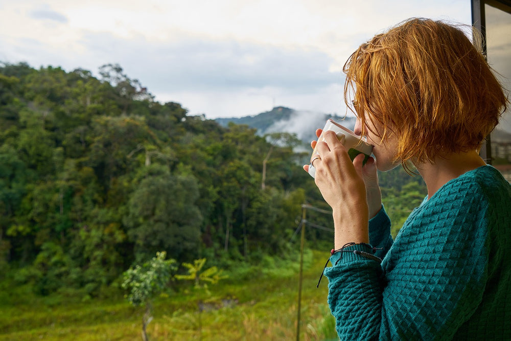 mujer con pelo rojo y abrigo verde que toma una taza de infusion mientras mira una valle con una montagña